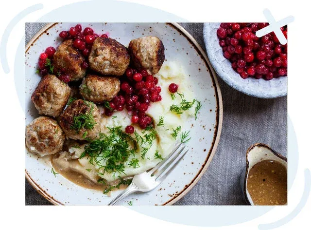 Plate of Scandinavian-style meatballs with mashed potatoes, gravy, lingonberries, and fresh dill, with a side bowl of berries and a small pitcher of sauce.