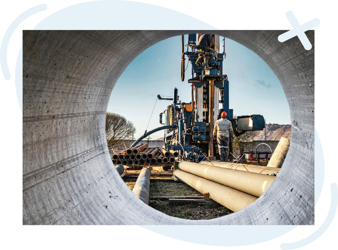 View through a large concrete pipe at a construction site with drilling machinery, stacked pipes, and a worker in a hard hat.