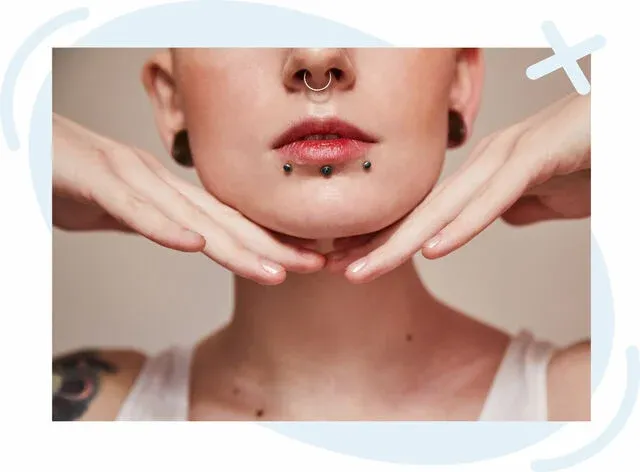 Close-up of a person's lower face with multiple facial piercings and hands framing the chin.