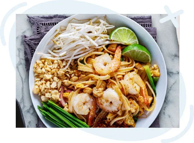 Top-down view of a plate of shrimp pad thai garnished with lime wedges, bean sprouts, crushed peanuts, and chives.