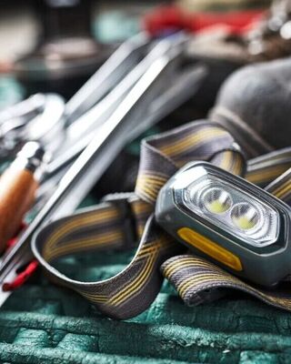 Close-up of camping gear on a green sleeping pad, featuring a gray headlamp with a striped elastic strap in the foreground, alongside metal tent stakes with wooden-handled mallet and red cords, and a rugged hiking boot in the background.