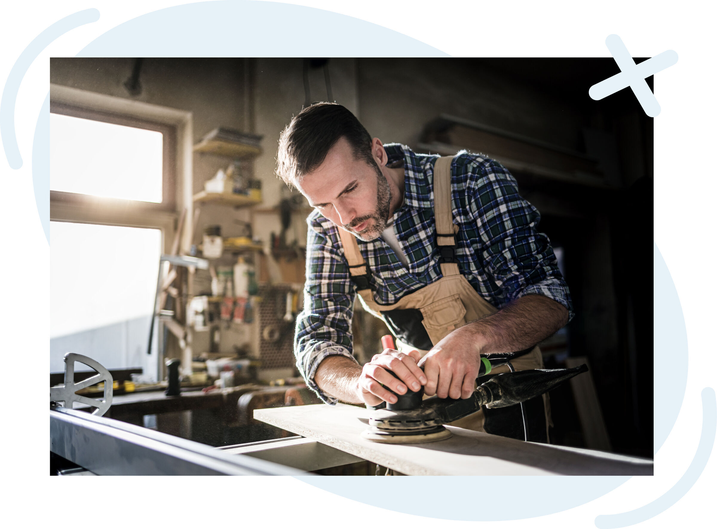 Carpenter using an electric sander on a wooden board in a sunlit workshop.