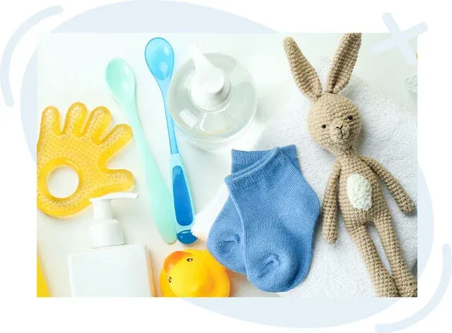 Flat lay of baby care items including a crocheted bunny toy, blue infant socks, teething ring, spoons, rubber duck, pump bottle, and towel on a white background.