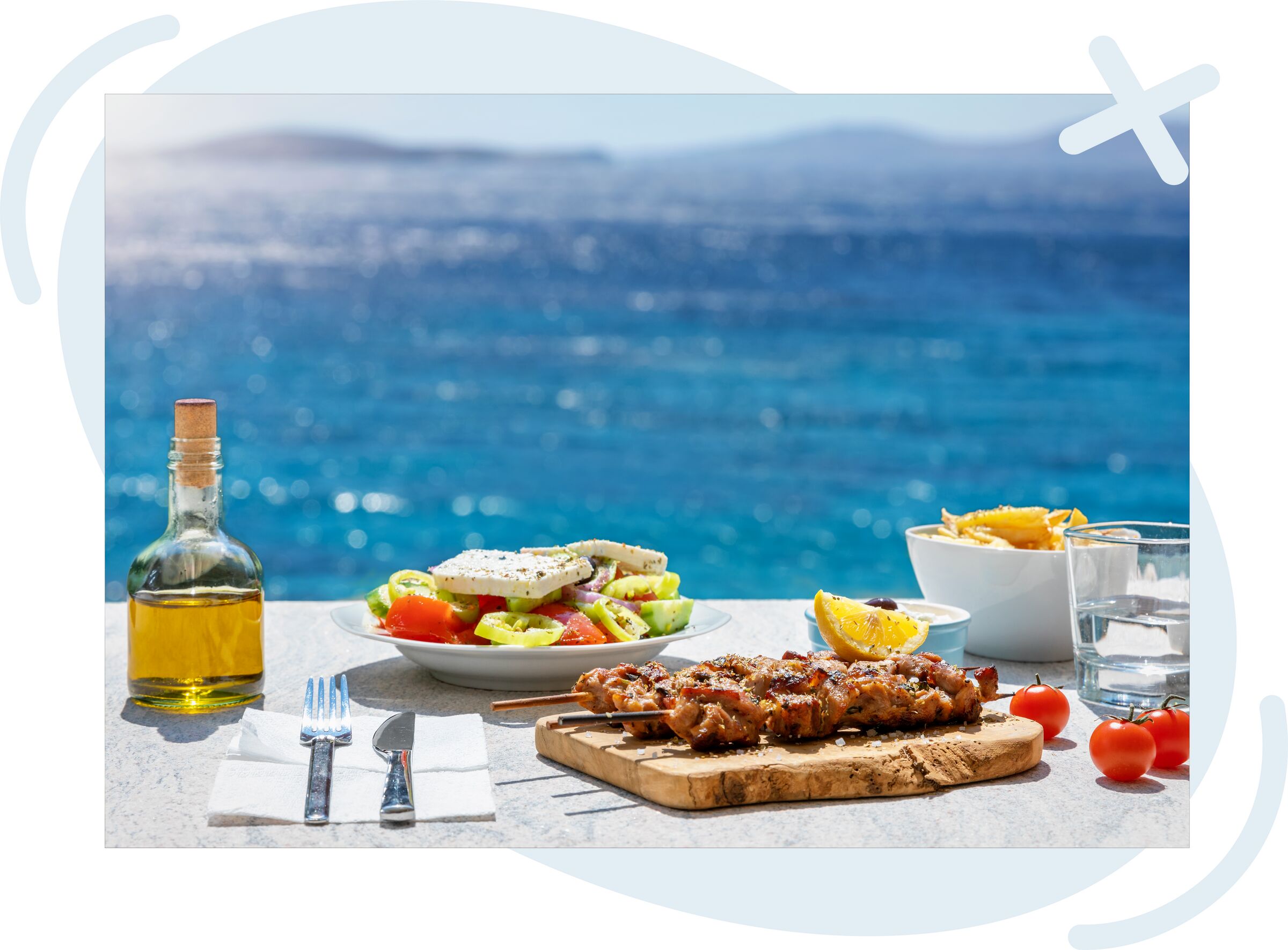Seaside outdoor meal with grilled skewers, Greek salad, fries, olive oil, and water on a sunlit table overlooking blue ocean.