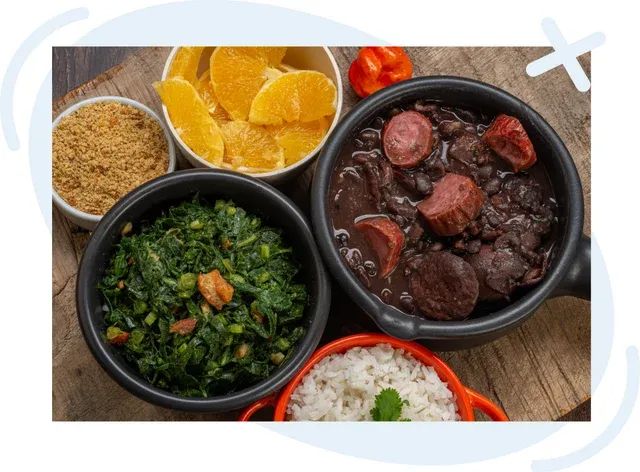 Traditional Brazilian meal with feijoada, collard greens, rice, orange slices, farofa, and a pepper on a rustic wooden table.